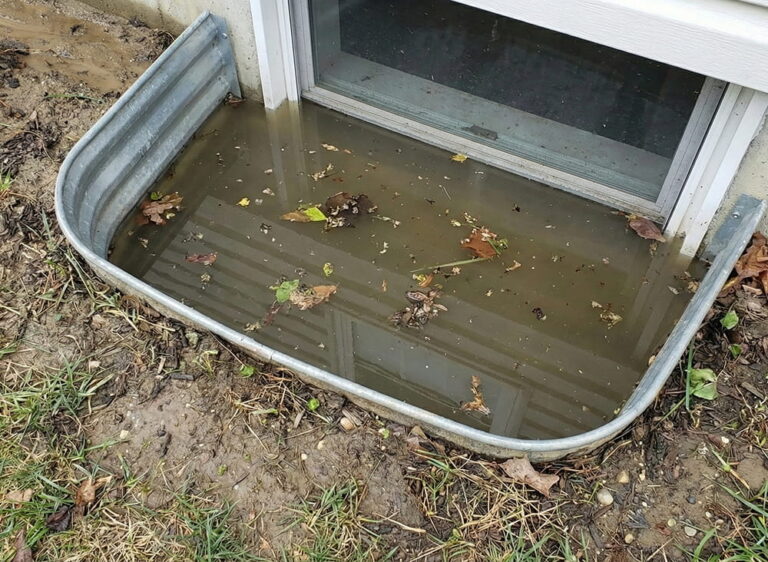 A corrugated metal basement window well completely filled with murky, standing water and debris due to poor surrounding grading.