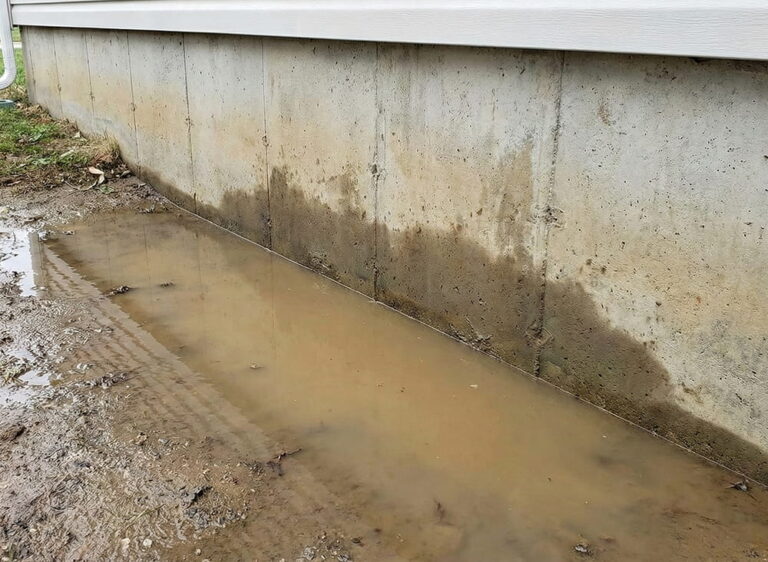 A large puddle of muddy water pooling directly against a concrete house foundation due to negative ground slope and poor drainage.